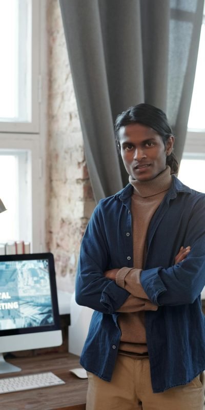 Young man posing confidently in a contemporary home office setting with digital marketing displayed on the computer screen.